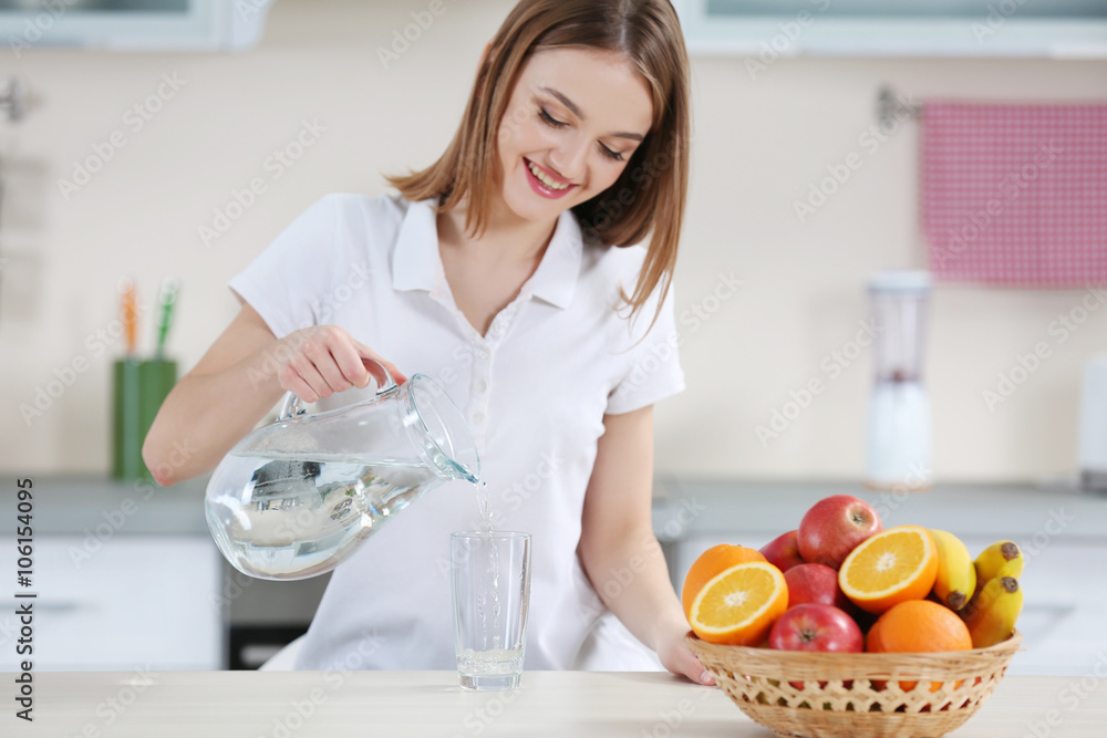 Young woman pouring water from jug into glass in the kitchen Young woman pouring water from jug into glass in the kitchen