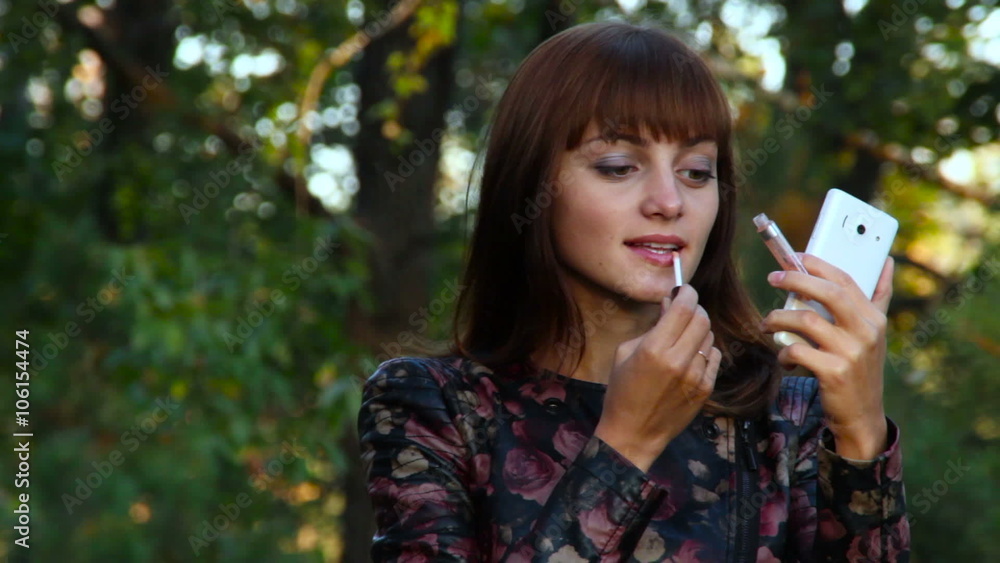 Pretty woman applying lip gloss looking at the smartphone Pretty woman applying lip gloss looking at the smartphone
