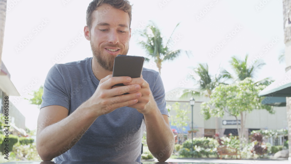 Handsome man using smartphone while sitting at sidewalk cafe. Smiling bearded male is text messaging on mobile cell smart phone. Young man is in casual clothing. RED EPIC. Handsome man using smartphone while sitting at sidewalk cafe. Smiling bearded male is text messaging on mobile cell smart phone. Young man is in casual clothing. RED EPIC.