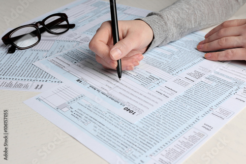 Female hand holding a pen next to the black rimmed glasses and filling in the 1040 Individual Income Tax Return Form for 2015 year on the white desk, close up
