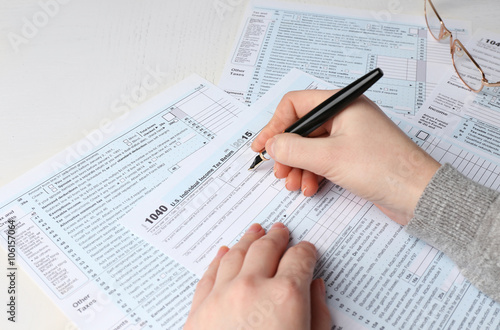 Female hand holding a pen next to the metal rimmed glasses and filling in the 1040 Individual Income Tax Return Form for 2015 year on the white desk  close up