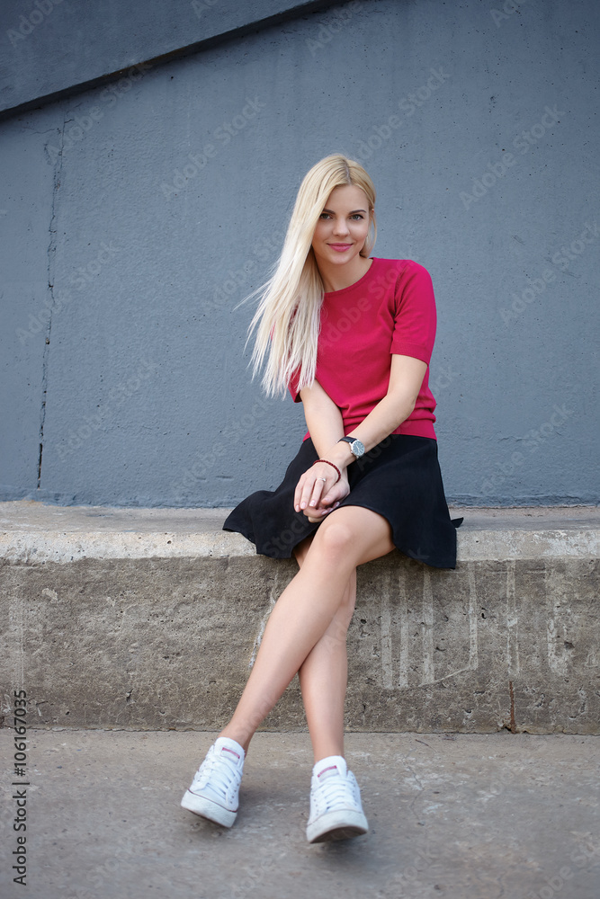 Young happy beautiful blonde woman sitting on the concrete river pier and looking into camera