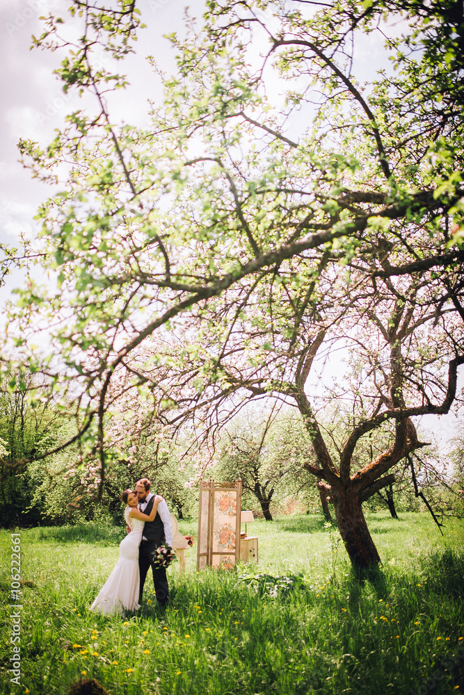 Elegant stylish groom lovely embraces with his happy gorgeous brunette bride on the background of trees in the park. Groom with beard
