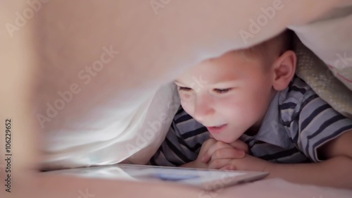 Boy lying under blanket and watching cartoon on touch pad