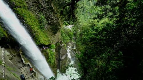 A high-angle shot of the Pailon del Diablo waterfall cascading down a moss-covered cliff in dense green forest, visitors on stone pathways and viewing platforms below near Banos, Ecuador.