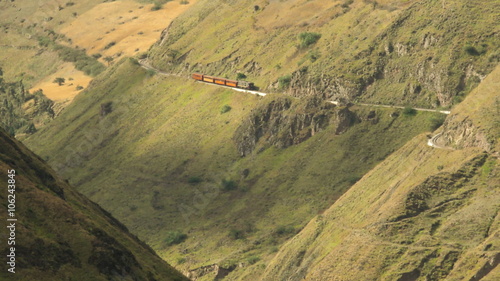 An aerial view of a red train moving along a narrow track carved into a steep green mountain valley on the Nariz del Diablo route near Alausi in the Ecuadorian Andes.
