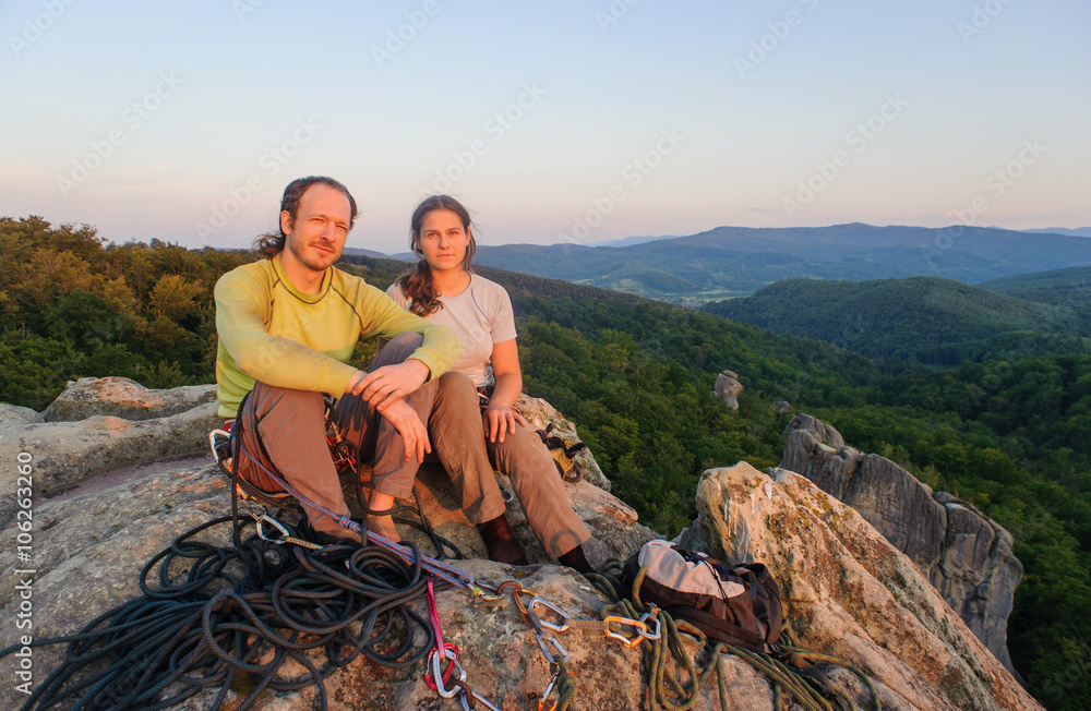 Young couple of climbers resting and enjoying beautiful nature view at sunset on the top of the mountain after challenging climb. Climbing equipment.