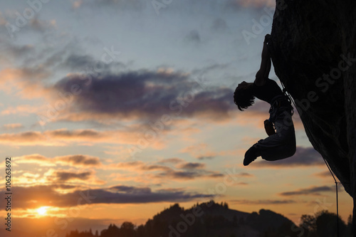 Silhouette of strong athletic female rock climber hanging on the natural rock cliff against beautiful sunset sky in the mountains. Summer time.