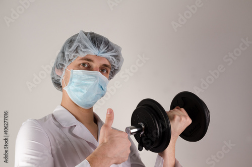 young man doctor keep dambbell in a hand and show like on neutral background. Medcine and sport concept. photo