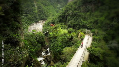 A high-angle view of two hikers crossing a wooden suspension bridge over a deep verdant gorge, a winding river and dense tropical forest below at the Pailon del Diablo near Banos, Ecuador.