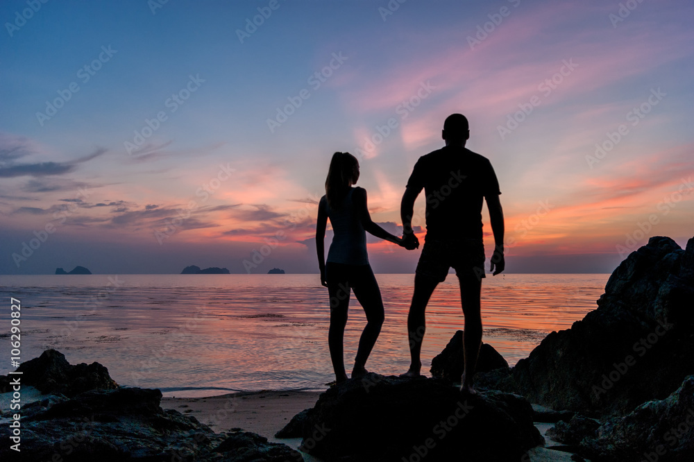 Silhouette young couple standing on the beach holding hands and watching  the tropical sunset 

