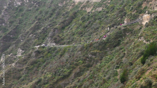 An aerial tracking shot of a winding mountain trail with hikers ascending a rugged green cliffside of the Quilotoa crater in the Ecuadorian Andes, revealing the turquoise lake below.