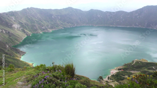 A panoramic high-angle shot of the Quilotoa crater lake surrounded by lush green volcanic slopes in the Ecuadorian Andes, wildflowers in the foreground and mist hovering over the turquoise water.