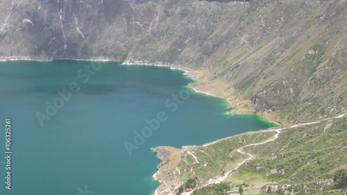 Aerial drone footage over the turquoise crater lake of Quilotoa in the Ecuadorian Andes, from a wide mountain vista to a closer view of the shoreline, a small beach and winding paths.