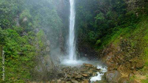 A powerful waterfall plunges down a moss-covered cliff into a rocky pool ringed by dense jungle and mist in the Ecuadorian rainforest near Banos.