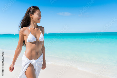 Bikini woman relaxing in white sun protection beachwear walking on tropical Caribbean beach with turquoise ocean water during summer vacations. Happy lifestyle Asian girl.