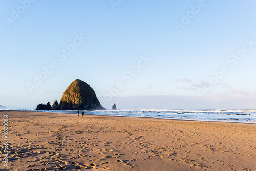 people walking in front of the haystack rock and the needles at cannon beach on the west coast of washington state in the united states of america at the sunrise with prints, wave and ocean