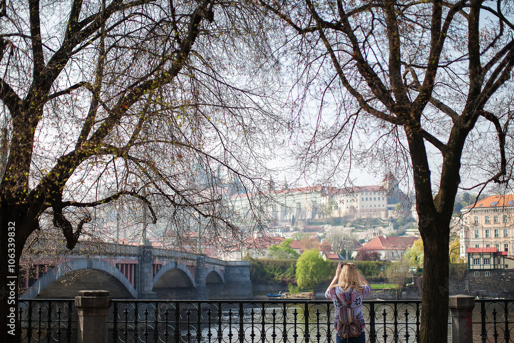 woman photographing city of Prague while standing on the waterfront among the trees