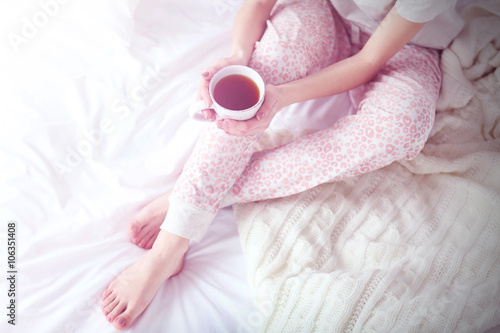 Woman in a pajamas holding a cup of tea and sitting in white bedding and pillows