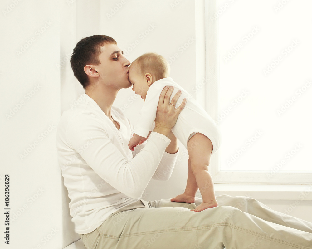 Happy young father kissing baby at home in white room near windo Happy young father kissing baby at home in white room near windo