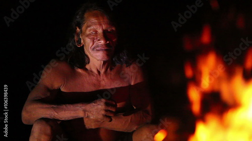 Editorial: an elderly Huaorani man sits by a roaring campfire at night, his face lit by the flames as he holds a wooden object, in the Yasuni reserve of the Ecuadorian Amazon.