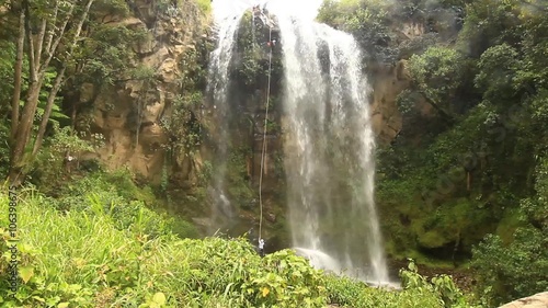 A person rappels down a tall cascading waterfall surrounded by dense green jungle, shot from a wide elevated perspective in the Ecuadorian rainforest near Banos.
