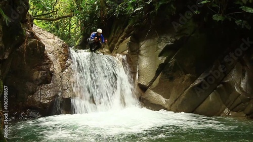A person in outdoor gear and helmet stands atop a small cascading waterfall in lush green jungle, surrounded by moss-covered rocks and dense foliage near Banos, Ecuador.