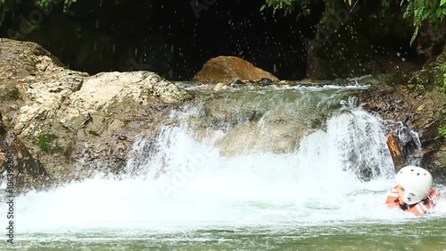 A group of adventurers in safety gear wade through a rushing jungle river and a small cascading waterfall, smiling at the camera amid lush greenery near Banos, Ecuador.