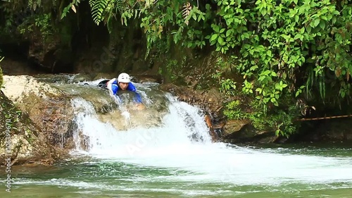 A kayaker in a blue jacket and white helmet runs a small rocky waterfall on a lush jungle river near Banos, Ecuador, emerging into calmer water amid dense green foliage.