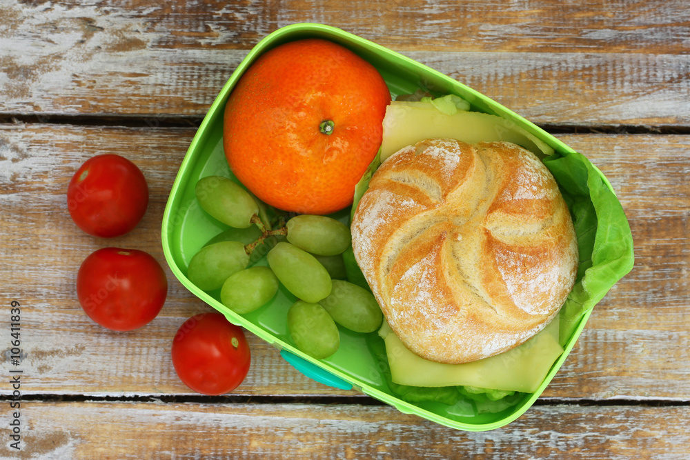 Lunch box containing cheese sandwich, mandarine, grapes and cherry tomatoes on wooden surface
