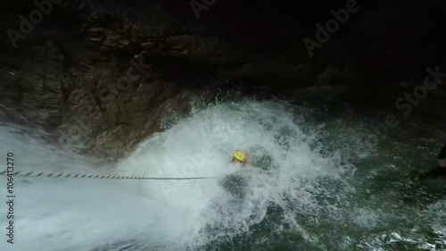 A person in a wetsuit, helmet and life vest rappels down a cascading waterfall inside a dark cave into turbulent white water, secured by a rope, near Banos, Ecuador.