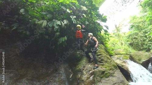 A person in safety gear jumps from a mossy rock into a rushing waterfall pool in lush jungle near Banos, Ecuador, a guide assisting, the camera following the jump and splash.