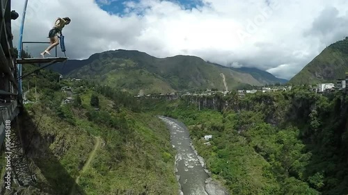 A person prepares to bungee jump from a high platform over a deep lush canyon with a winding river, distant mountains and buildings below, at the San Francisco bridge near Banos, Ecuador.