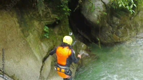 A person in a yellow helmet and orange life vest jumps from a mossy rock into a rushing river creating a splash as another watches from above in lush jungle, canyoning near Banos, Ecuador.