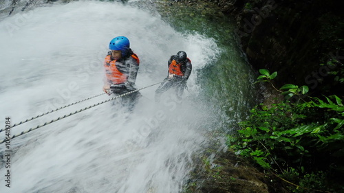 Two adventurers in safety gear rappel down a cascading waterfall in a lush rocky canyon, descending ropes through rushing water and moss-covered rocks, canyoning near Banos, Ecuador.