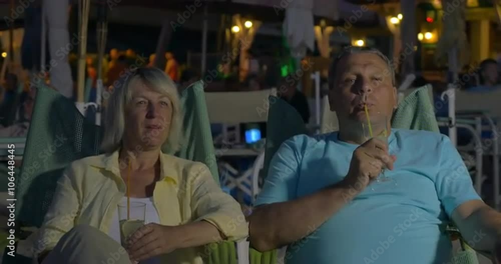 Happy senior family couple spending evening on the beach. They sitting in chaise-longues, having drinks and enjoying summer vacation Happy senior family couple spending evening on the beach. They sitting in chaise-longues, having drinks and enjoying summer vacation
