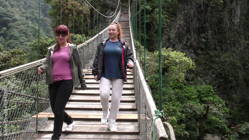 Two women walk across a wooden suspension bridge in a lush jungle canyon near Banos, Ecuador, the camera panning right to reveal a tall waterfall cascading down a rocky cliffside.
