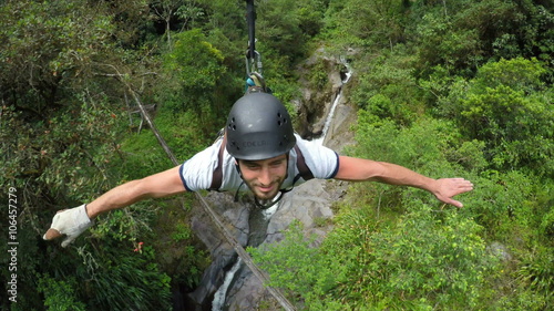 A man in a helmet and harness ziplines through dense tropical jungle near Banos, Ecuador, arms outstretched with a waterfall and rocky terrain below, a high-angle tracking shot.