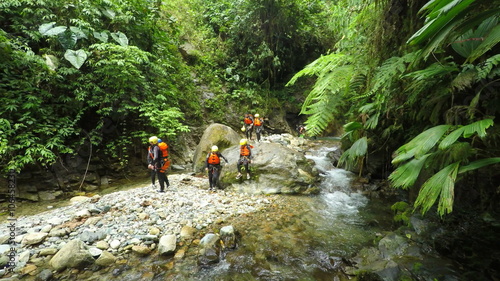 A group of adventurers in helmets and life vests cross a rocky stream over boulders in lush tropical jungle of Cashaurco Canyon near Banos, Ecuador, amid dense green foliage and flowing water.