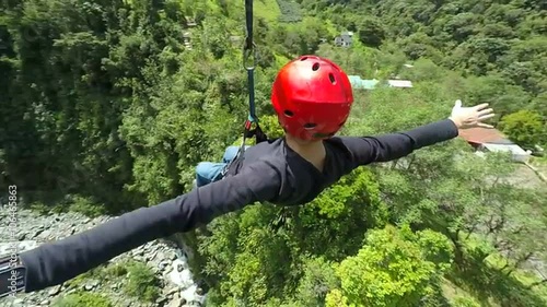 A man in a red helmet and harness ziplines across a deep forested valley with arms outstretched, a rocky riverbed and distant mountains below near Banos, Ecuador, high angle.