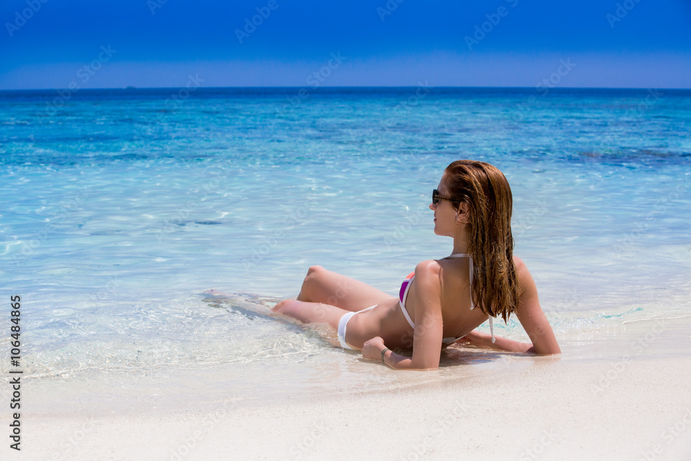 Sexy woman in bikini lying and sunbathing on tropical beach. Slim girl with long legs and smooth skin relaxing at the water edge.  Beautiful beach with white sand and azure ocean.