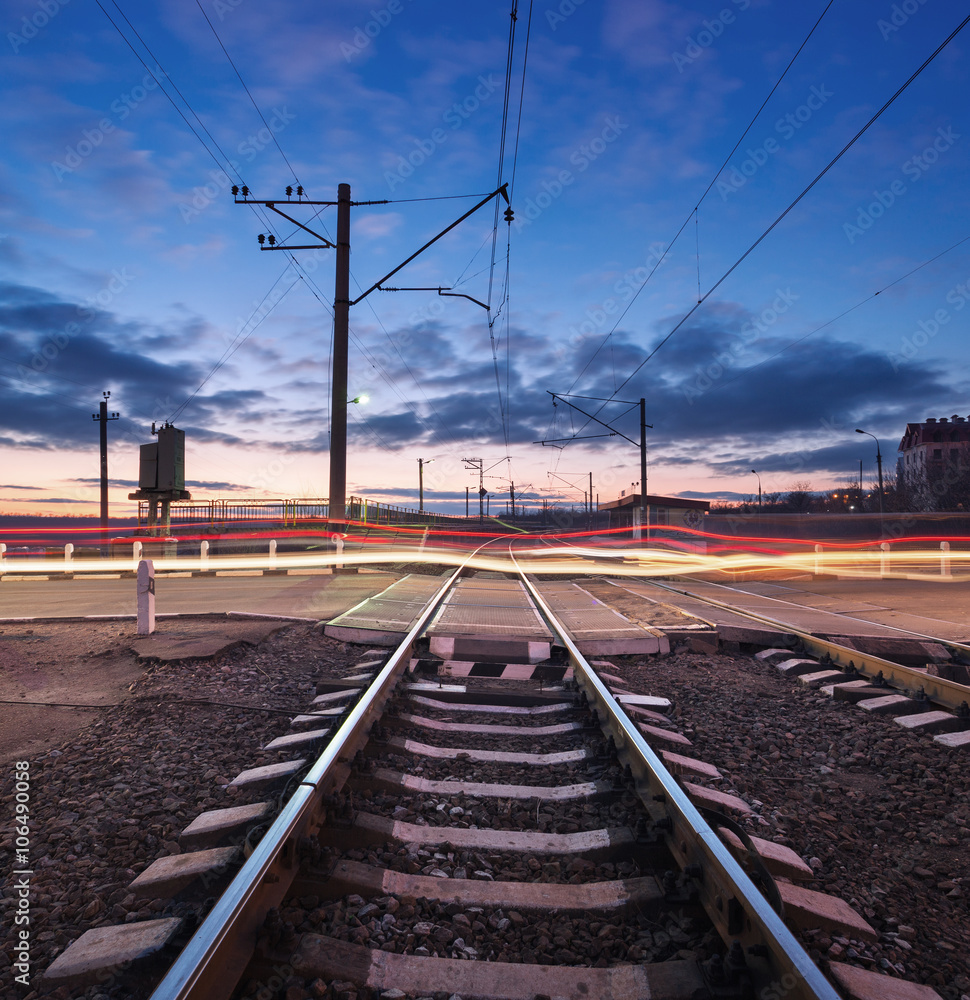Rail crossing with blurred car lights on the background of colorful cloudy sky at beautiful sunset. Railway landscape