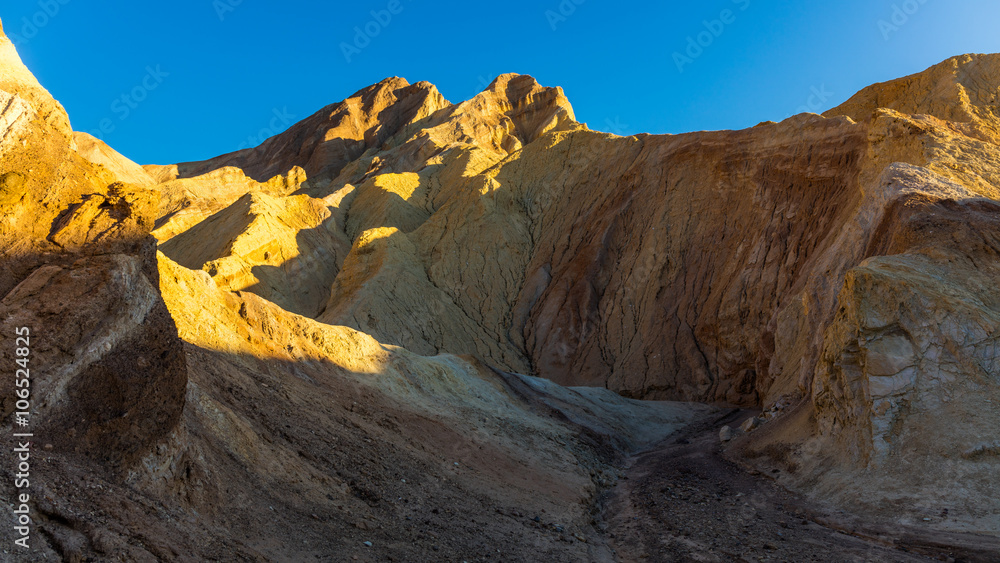 High sandstone cliffs painted in many glowing shades of orange, gold and red. Narrow canyon with vertical walls on both sides. Rocky landscape background. Golden canyon, Death Valley
