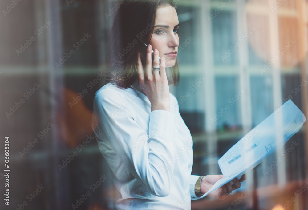 Photo business woman wearing white shirt, talking smartphone and holding documents in hands. Open space loft office. Panoramic windows background. Horizontal mockup, film effect Photo business woman wearing white shirt, talking smartphone and holding documents in hands. Open space loft office. Panoramic windows background. Horizontal mockup, film effect