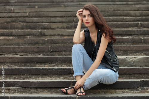 Young thoughtful fashionable brunette woman in black leather jacket posing sitting on park stairway