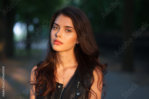 Closeup portrait of happy young beautiful brunette woman in black leather jacket posing on sunset outdoors with blurry foliage background