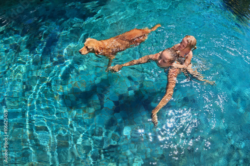 Mother with little child on back play with fun and train golden labrador retriever puppy in swimming pool. Popular dog like companion, outdoor activity and funny game with family pet on summer holiday
