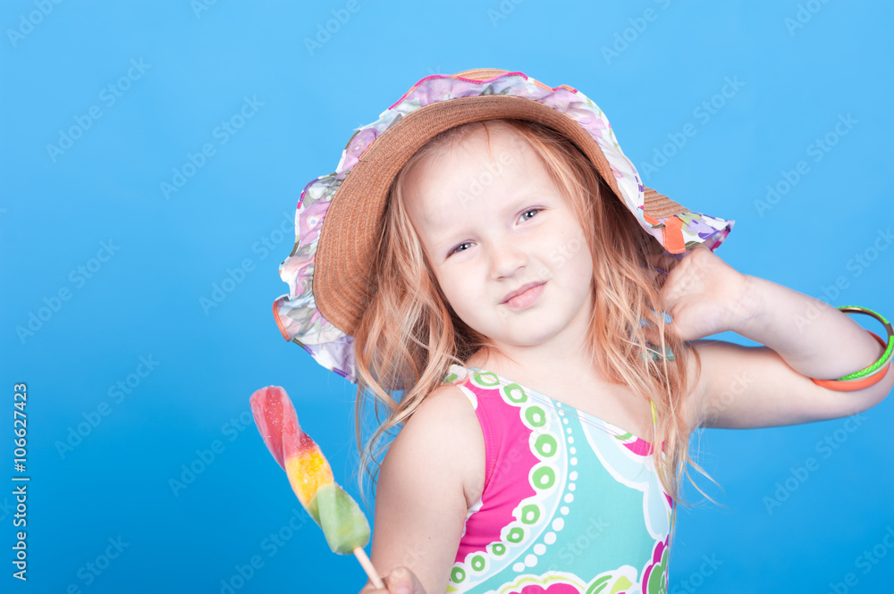 Smiling baby girl 4-5 year old eating ice cream in room over blue. Wearing hat and swim suit. Looking at camera. Smiling baby girl 4-5 year old eating ice cream in room over blue. Wearing hat and swim suit. Looking at camera.