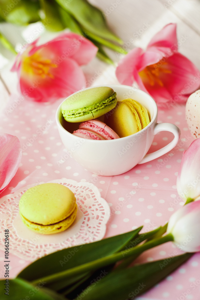 A beautiful flowers pink tulips with colorful macaroons laid in cup on white wooden background with pink Lacy napkin A beautiful flowers pink tulips with colorful macaroons laid in cup on white wooden background with pink Lacy napkin
