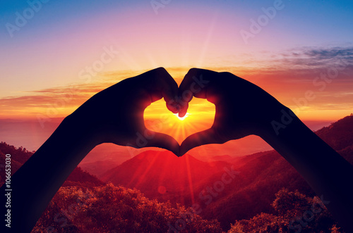 Valentine's day concept, Silhouette (Focused) hand in heart shape with sunset in the middle and mountain (Blurred) background